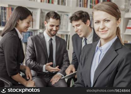 Business woman smiling and looking at the camera with her colleagues talking and looking down at a digital tablet in the background