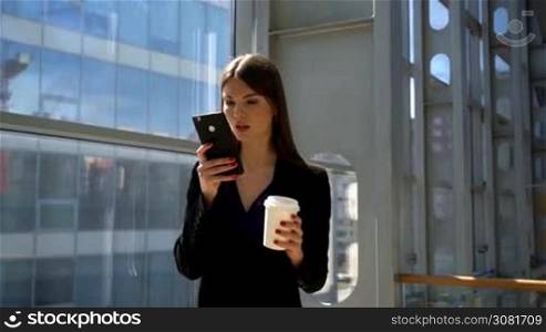 Business woman walking in business center holding coffee and surfing the net at mobile phone
