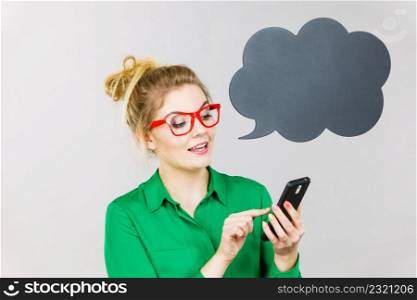 Business woman wearing green shirt and red eyeglasses looking at phone with black thinking or speech bubble next to her.. Business woman looking at phone, thinking bubble