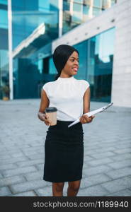 Business woman with cardboard coffee cup and notepad outdoors, office building on background. Black businesswoman in skirt and white blouse. Business woman with coffee cup and notepad