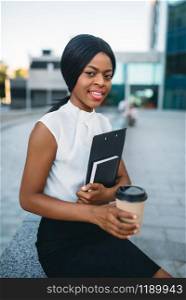 Business woman with cardboard coffee cup and notepad resting during lunch break outdoors, office building on background. Black businesswoman in skirt and white blouse