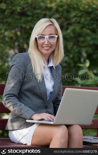 Business woman with computer laptop looking at camera in business district city park, sitting on bench