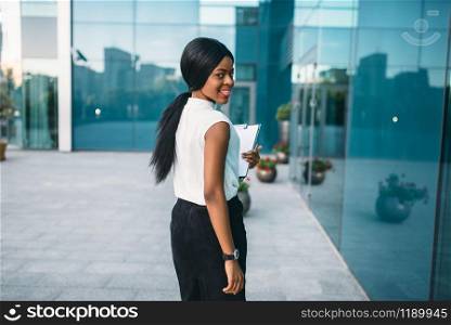 Business woman with notepad outdoors, back view, office building on background. Black businesswoman in skirt and white blouse. Business woman with notepad outdoors, back view