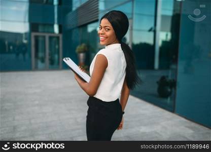 Business woman with notepad outdoors, back view, office building on background. Black businesswoman in skirt and white blouse