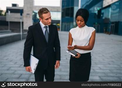 Business woman with phone and businessman with laptop in hands walking outdoors, modern office building on background, partnership negotiations during the lunch break. Successful businesspeople. Business woman with phone, businessman with laptop
