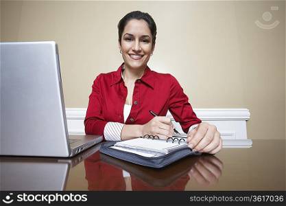 Business woman writing in diary beside laptop in office, portrait