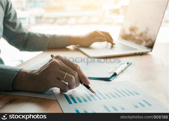 Businessman analyzing annual business report and holding pen and pointing paper chart summary with using laptop at room office desk.