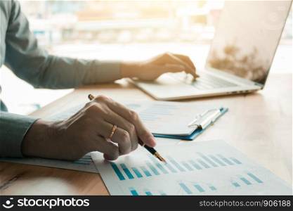Businessman analyzing annual business report and holding pen and pointing paper chart summary with using laptop at room office desk.