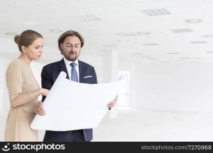 Businessman and businesswoman looking at blueprint while standing in new empty office