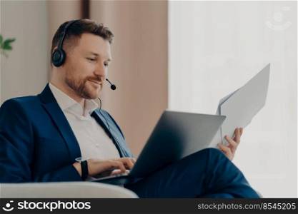 Businessman at work. Smiling bearded male boss holding documents and laptop on his lap while sitting indoors and having an online meeting. Business people and modern technologies. Thoughtful businessman working with online project at home while holding papers