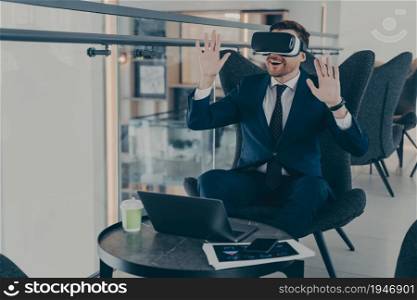 Businessman dressed in formal suit wearing portable VR goggles exploring virtual world while sitting on comfortable chair in cafe and working remotely, expressing positive emotions with his gestures. Businessman dressed in formal suit wearing VR goggles while working on laptop in cafe