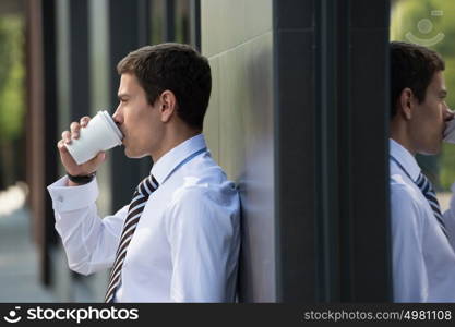 Businessman drinking coffee leaning on wall of modern office building. Urban professional smiling happy wearing white shirt holding disposable coffee cup. Handsome male model in his twenties.