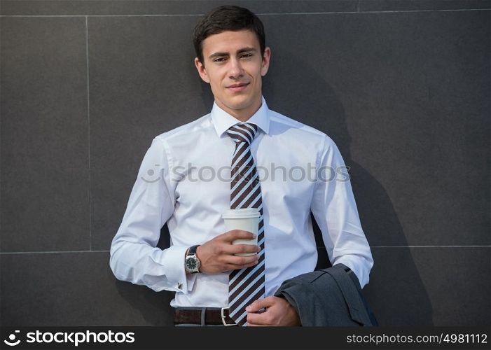 Businessman drinking coffee leaning on wall of modern office building. Urban professional smiling happy wearing white shirt holding disposable coffee cup. Handsome male model in his twenties.