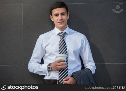 Businessman drinking coffee leaning on wall of modern office building. Urban professional smiling happy wearing white shirt holding disposable coffee cup. Handsome male model in his twenties.