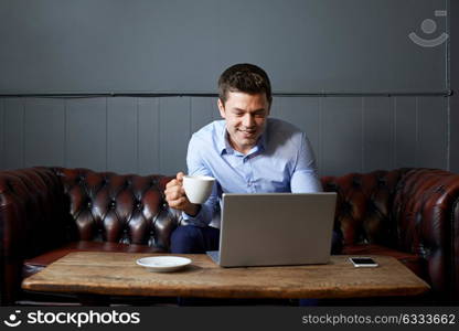 Businessman Drinking Coffee Whilst Working On Laptop In Internet Cafe