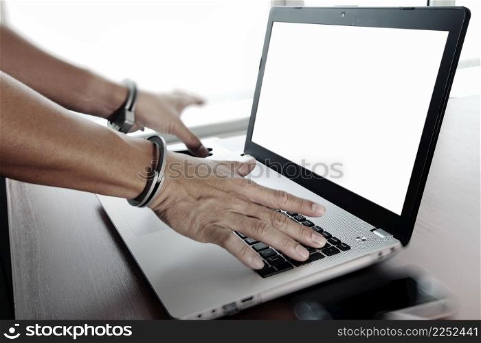 Businessman hand in handcuffs at wooden desk with laptop computer and digital tablet and stylus pen and smart phone as Cyber-Crime concept