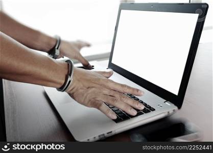 Businessman hand in handcuffs at wooden desk with laptop computer and digital tablet and stylus pen and smart phone as Cyber-Crime concept