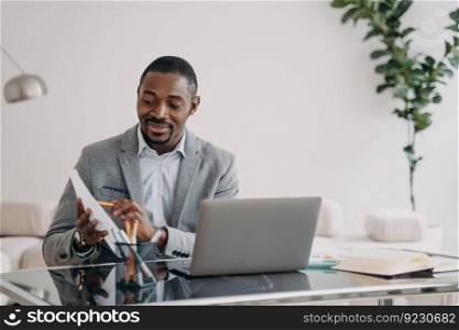 Businessman has paperwork. Happy man has video call on laptop and pointing to data papers with pencil. Freelancer is working online at home office. Remote african american worker.. Businessman has paperwork. Happy man has video call on laptop and pointing to data papers.