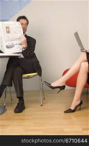 Businessman holding a newspaper and looking at the legs of a businesswoman working on a laptop