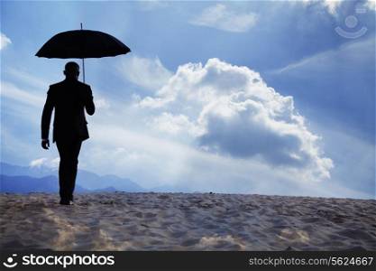 Businessman holding an umbrella and walking away in the middle of the desert with dreamlike clouds