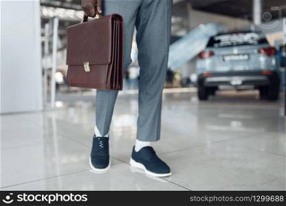 Businessman holds briefcase in car showroom. Successful business person on motor show, black man in formal wear