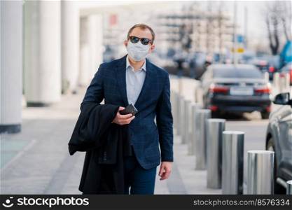 Businessman in black elegant clothes, medical mask, holds smartphone in hands, walks through busy city with many transport, protects himself from coronavirus during pandemic. Virus protection