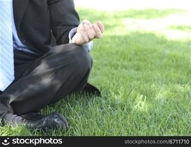 Businessman in black suit, blue shirt, and blue tie is sitting meditation style in the green grass