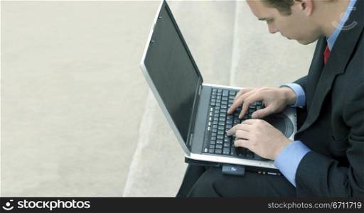 Businessman in blue shirt, red tie, and black suit is busily typing on his laptop on the steps of the courthouse
