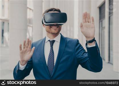 Businessman in blue suit trying out and experiencing virtual reality using mobile VR headset, planning to use it for advertising and future projects, standing alone in front of illuminated building. Businessman in suit trying out and experiencing virtual reality using mobile VR headset outdoors