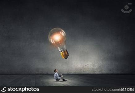 Businessman in concrete room. Young businessman sitting on floor and looking up on glowing bulb