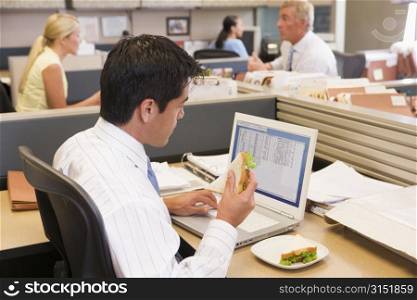Businessman in cubicle at laptop eating sandwich