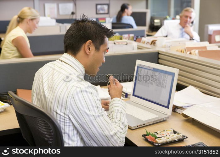 Businessman in cubicle at laptop eating sushi