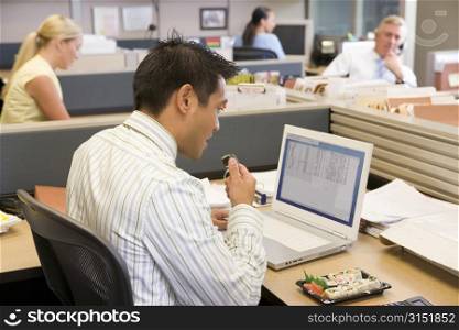 Businessman in cubicle at laptop eating sushi