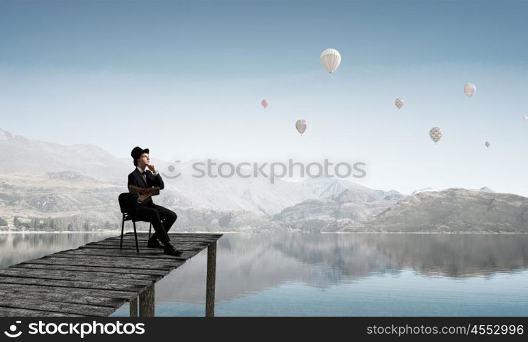 Businessman in cylinder reading book. Young businessman at pier sitting in chair with old book in hands