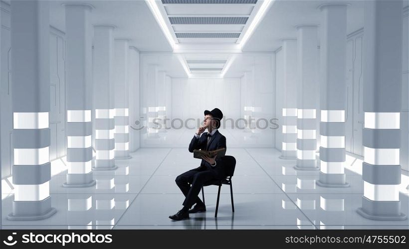 Businessman in cylinder reading book. Young businessman in futuristic interior sitting in chair with old book in hands