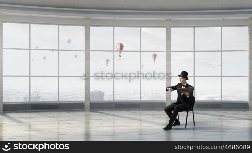 Businessman in cylinder reading book. Young businessman in office sitting in chair with old book in hands