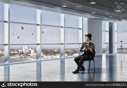 Businessman in cylinder reading book. Young businessman in office sitting in chair with old book in hands