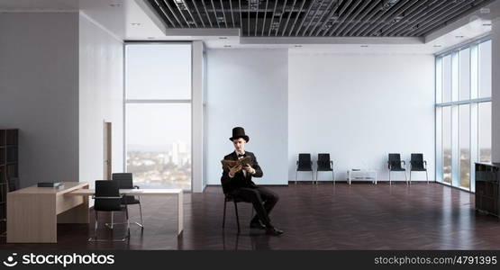 Businessman in cylinder reading book. Young businessman in office sitting in chair with old book in hands