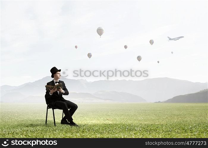 Businessman in cylinder reading book. Young businessman outdoor sitting in chair with old book in hands