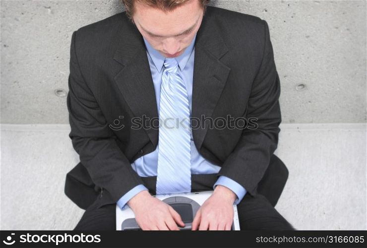 Businessman in dark blue shirt, blue tie, and blue suit is typing on his laptop against a concrete wall