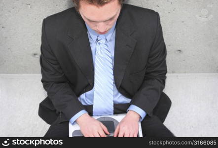 Businessman in dark blue shirt, blue tie, and blue suit is typing on his laptop against a concrete wall