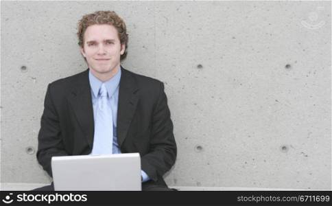 Businessman in dark blue suit, blue shirt, and blue tie is sitting up against concrete wall and typing on his laptop