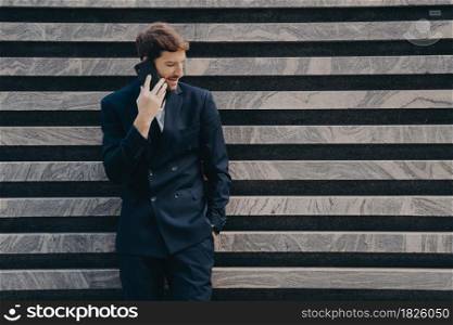 Businessman in elegant black clothes phones to partner focused down keeps smartphone near ear poses near marble wall copy space for your advertisement. People technology and bussines concept. Businessman in elegant black clothes phones to partner focused down keeps smartphone