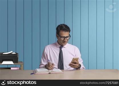 Businessman in eyeglasses writing in book while looking at his mobile phone sitting at his office desk