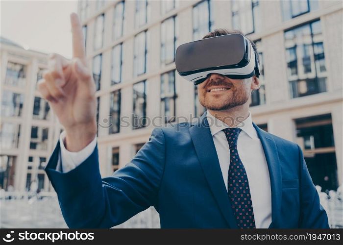 Businessman in formal blue suit using VR glasses outside, interacting with virtual world, trying to touch something in front of him while smiling, blurred office buildings on background. Businessman in formal blue suit using VR glasses outside