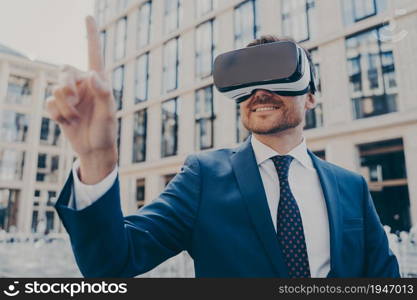 Businessman in formal blue suit using VR glasses outside, interacting with virtual world, trying to touch something in front of him while smiling, blurred office buildings on background. Businessman in formal blue suit using VR glasses outside