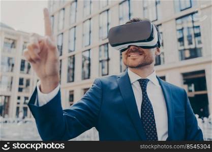 Businessman in formal blue suit using VR glasses outside, interacting with virtual world, trying to touch something in front of him while smiling, blurred office buildings on background. Businessman in formal blue suit using VR glasses outside