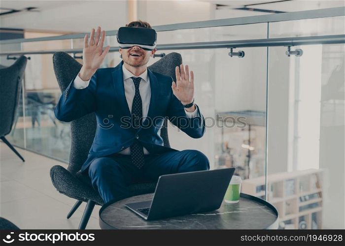 Businessman in formal suit and in VR headset glasses pointing with open palms up in air. Office worker or CEO immersed in 3D reality, innovative method of browsing web or managing business project. Businessman in formal suit and in VR headset glasses pointing with open palms up in air