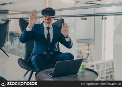 Businessman in formal suit and in VR headset glasses pointing with open palms up in air. Office worker or CEO immersed in 3D reality, innovative method of browsing web or managing business project. Businessman in formal suit and in VR headset glasses pointing with open palms up in air