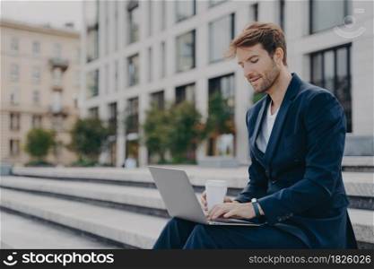 Businessman in formal suit reads news via laptop computer works online drinks takeaway coffee poses outdoors near office building sits on steps connected to wireless internet. Male entrepreneur. Businessman in formal suit reads news via laptop computer works online drinks takeaway coffee
